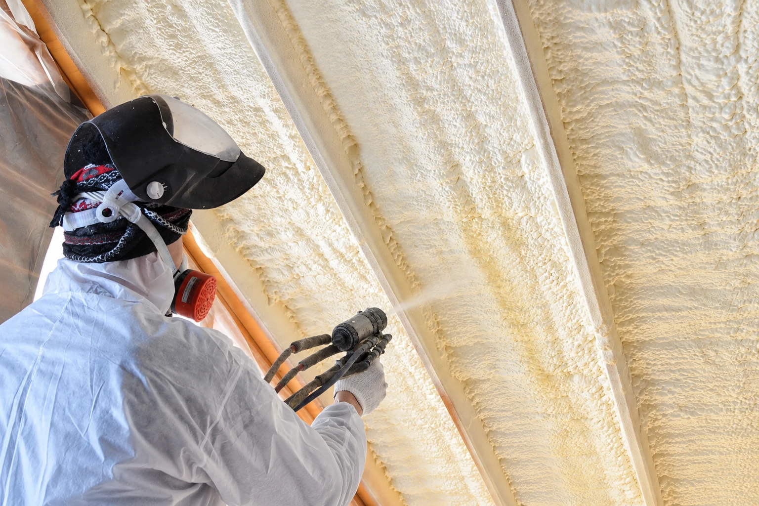 A worker in protective clothing sprays foam insulation onto the underside of a roof between wooden rafters.