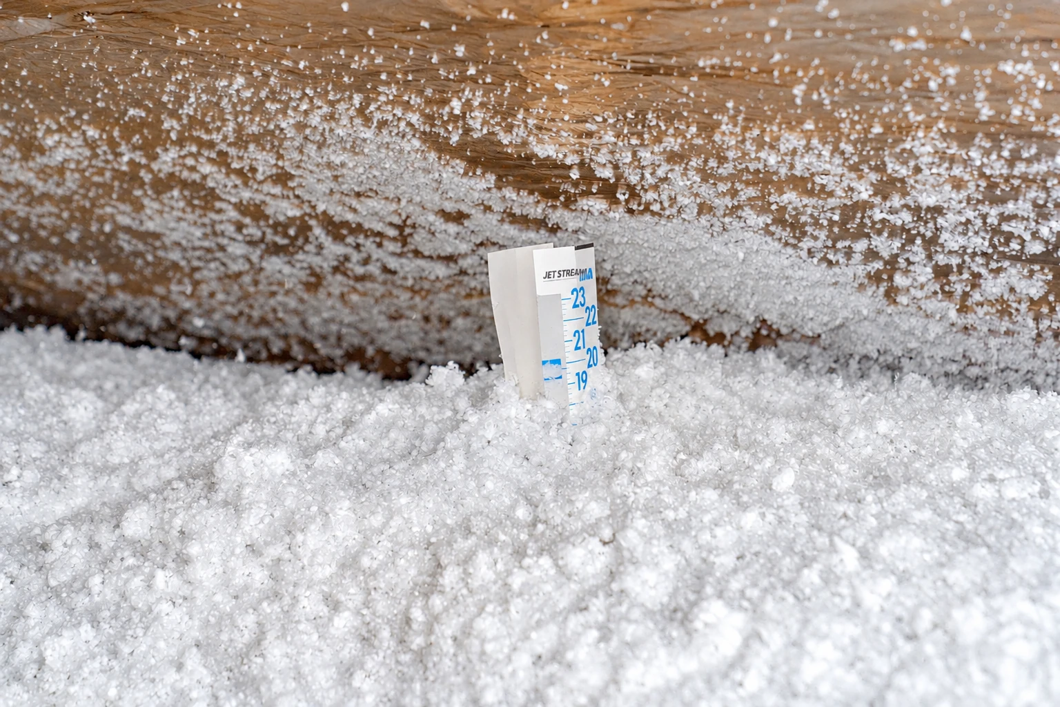 White blown-in insulation covers an attic floor, with a depth gauge standing upright in the material.