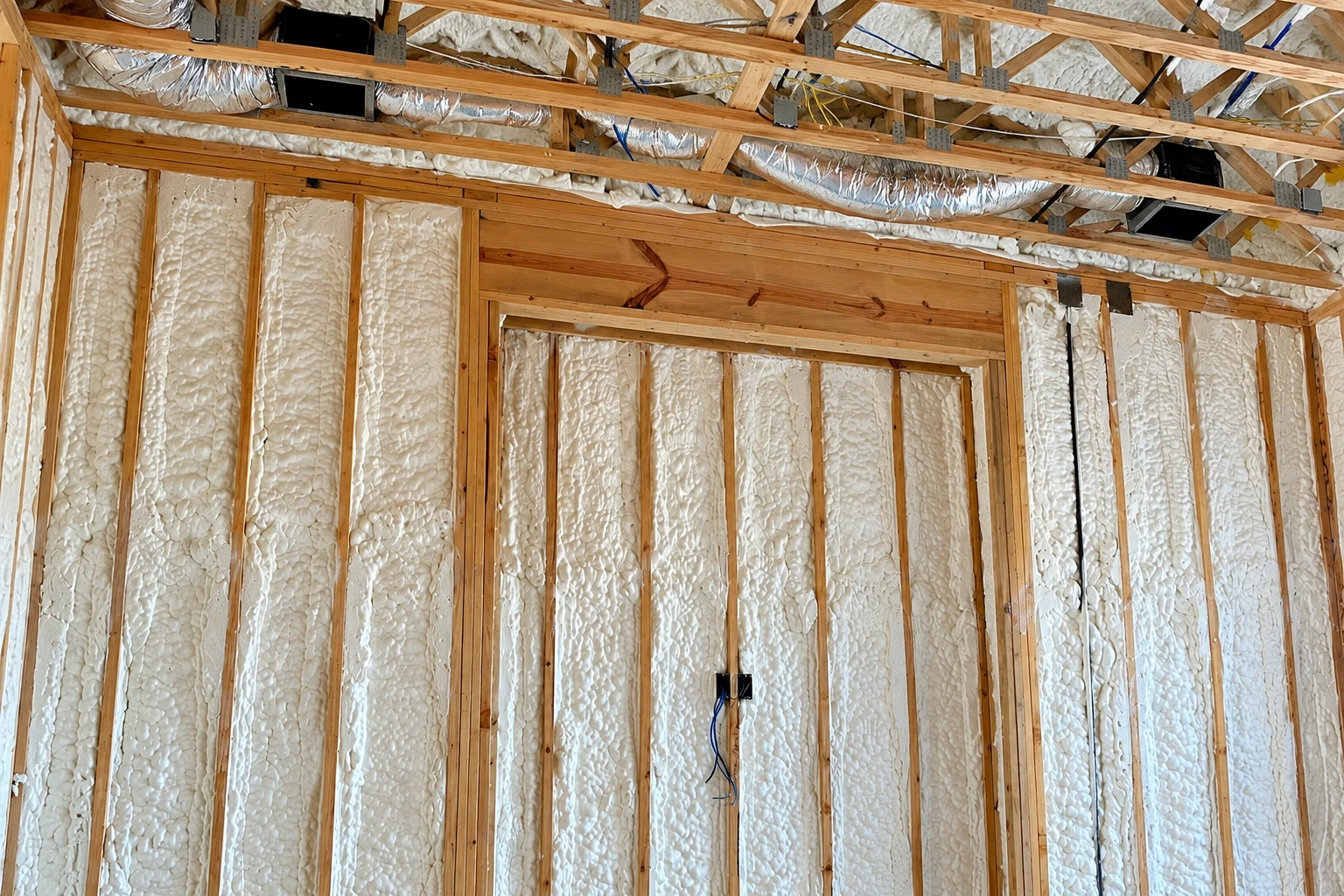 Exposed wall framing filled with white spray foam insulation beneath overhead ductwork.