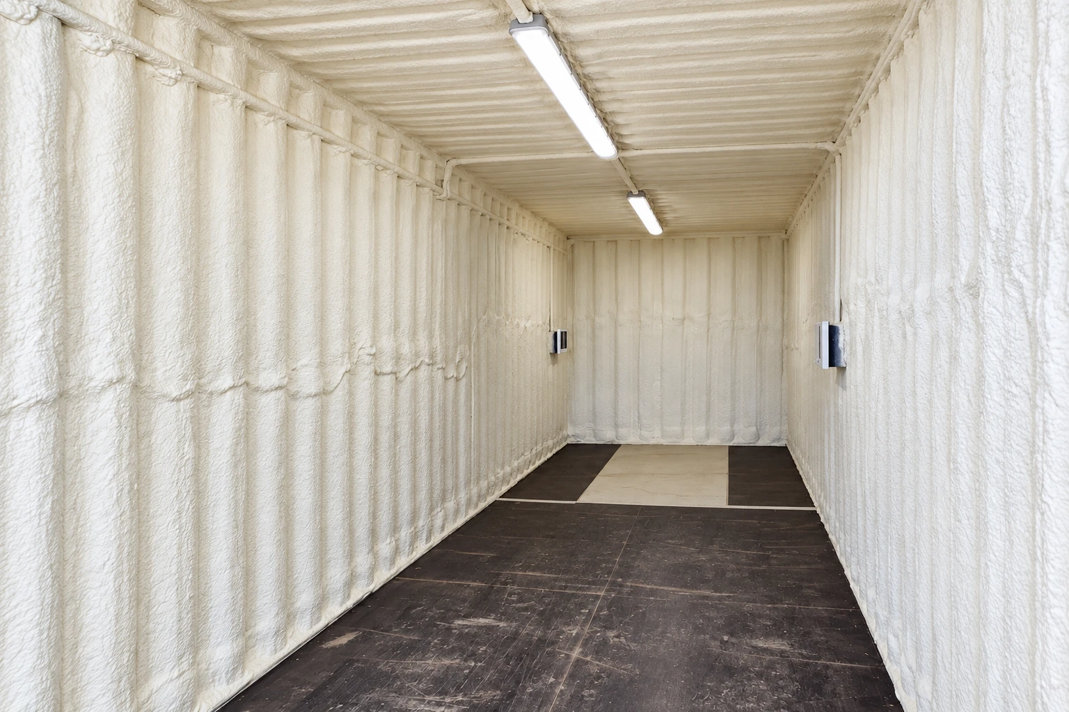 A long commercial container interior is coated with white spray foam on its walls and ceiling, with lights overhead.