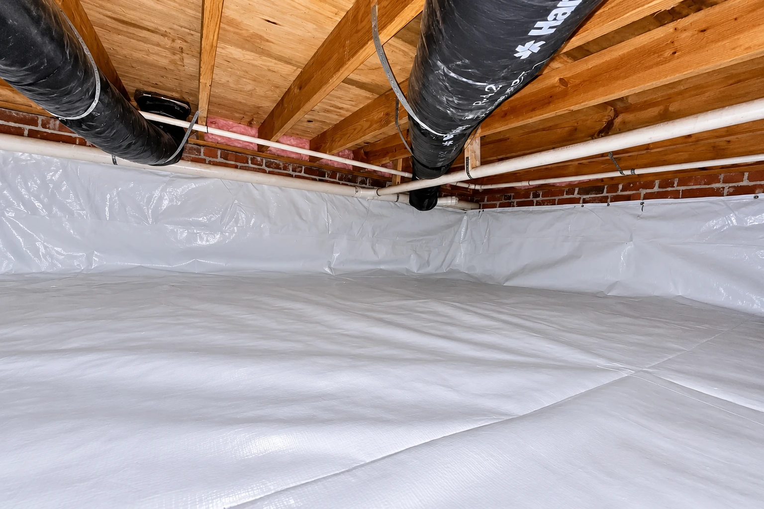 A crawl space has white vapor barrier sheeting across the floor and lower walls beneath ducts and pipes.
